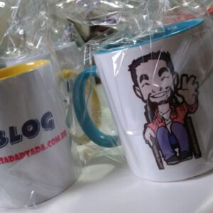 foto das canecas do blog dentro de sacos plásticos, uma com a logo do blog e outra mostrando o outro lado com o mascote tom, boneco, personagem do blog, homem cadeirante, com barba sorrindo e acenando com a mão esquerda, usa uma camisa de manga longa rosa, outra por dentro branca, calça jeans azul e sapatos pretos.