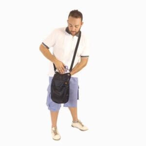 Imagem ilustrativa da bolsa na cor preta e detalhe laranja, na foto, um homem branco, cabelos curtos castanhos, barba, usa uma camisa branca com detalhes pretos e cinza nas mangas, short jeans azul, sapato na cor bege, o mesmo olha para baixo, inserindo um coletor de urina na bolsa.