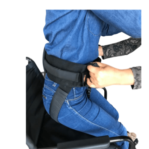 Foto ilustrativa em zoom da Cinta de transferência abdominal com alças laterais na cor preta, na imagem 2 modelos simulam o uso sentada na cadeira de rodas de cor preta se preparando para levantar, a primeira usa a cinta e um conjunto de blusa e calça jeans azul, atrás dela a segunda modelo de pele clara segura a cinta e a puxa, modelo usa uma camisa de manga longa com detalhes em cinza e preto.