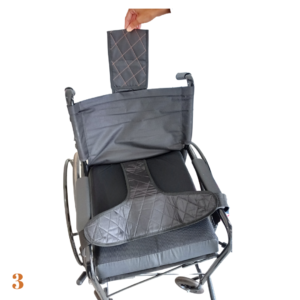 Foto ilustrativa do cinto de segurança pélvico como é encaixado na cadeira de rodas, na foto em questão mostra uma cadeira de rodas na cor cinza detalhes pretos, acima dA almofada e do encosto uma mão segura o produto mostrando o encaixe.